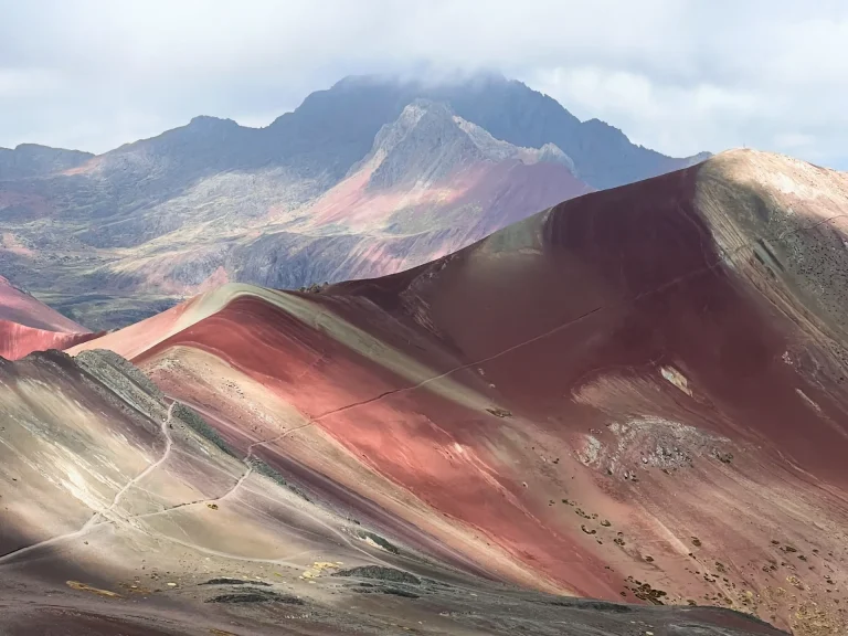 Spalvingi kalnai, padengti raudonų, rožinių ir žalsvų atspalvių juostomis, su ryškiu kalnų masyvu tolumoje. Šis vaizdas simbolizuoja gyvenimo kelio įvairiapusiškumą ir harmoniją, siejamą su Žmogaus Dizaino sistema. Human Design analizė padeda atrasti jūsų prigimtinį potencialą ir energijos stiprybę, kviečiant atrasti vidinę ramybę ir gyvenimo tikslą. Sužinokite daugiau apie save su GoToYourPower.lt.